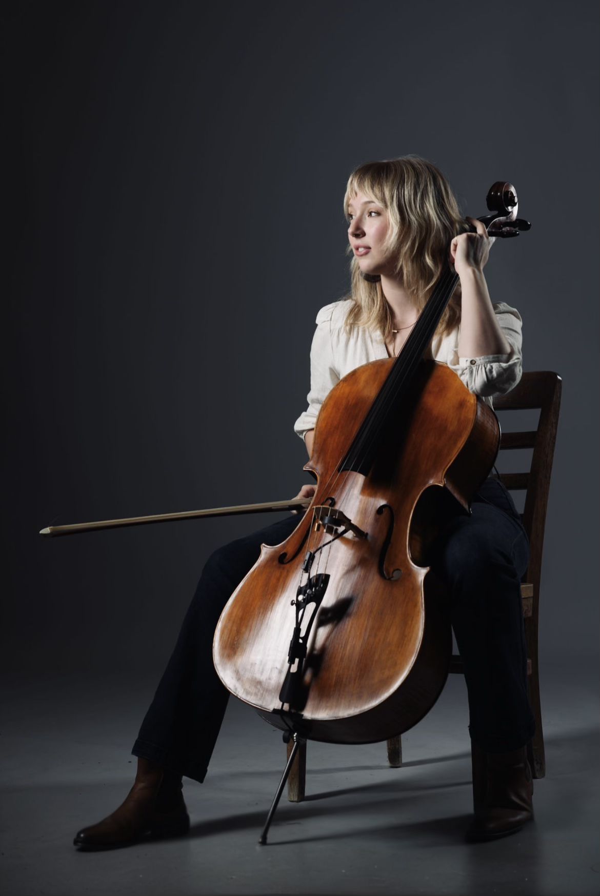 Annie Koppes seated with a cello, looking off-camera in a dark studio.
