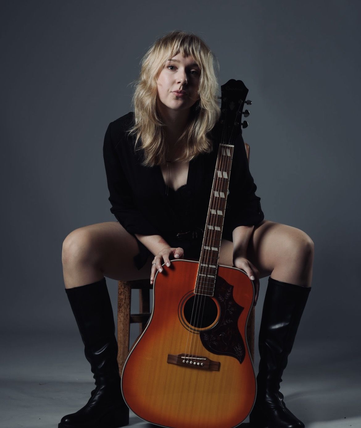 Annie Koppes seated with an acoustic guitar in a dark studio, looking at the camera.