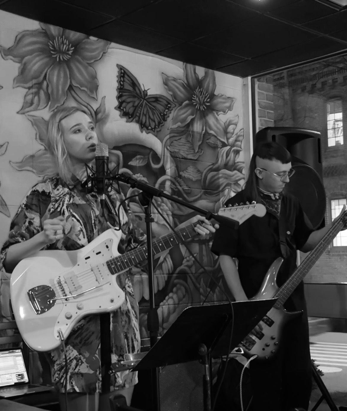 anneek performing live in black and white, singing into a condenser microphone with a Jazzmaster guitar, beside a bassist, in front of a painted floral mural.